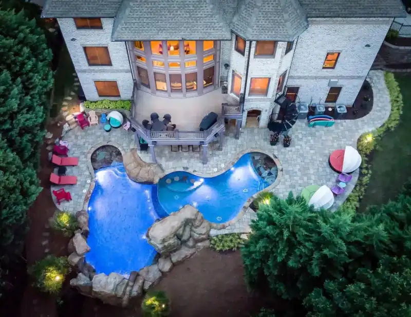 Night aerial of a lit pool with a rock grotto waterfall beside a white house